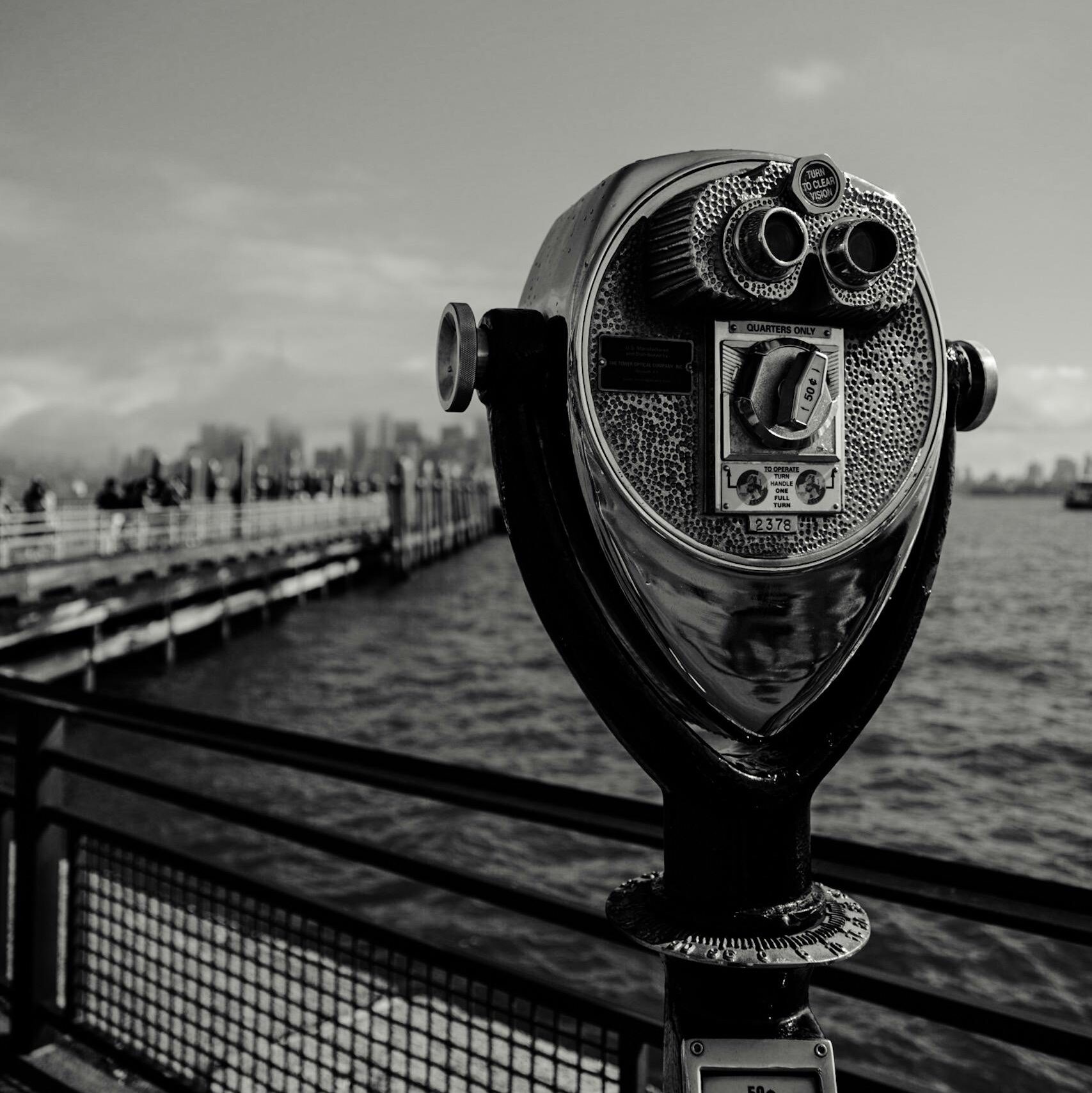 Black and white image of a coin-operated viewer on a pier overlooking the water.