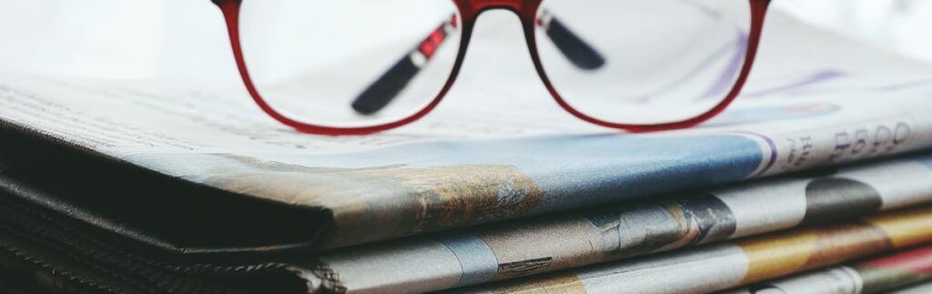 Red eyeglasses resting on a stack of newspapers symbolize reading and information.